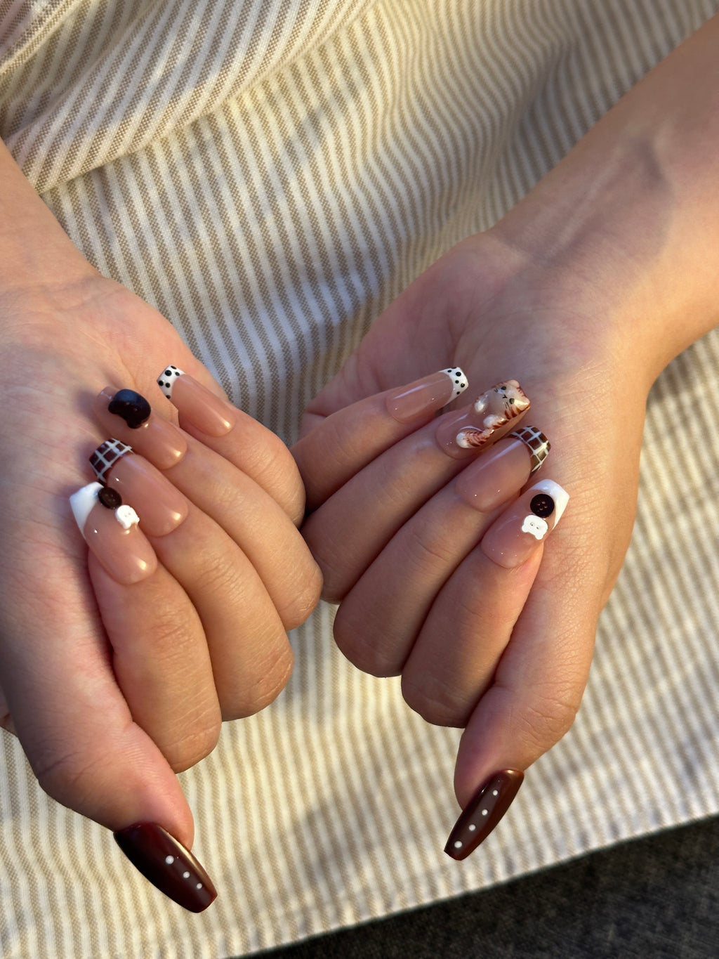 Both hands over a striped fabric background showcasing brown plaid French coffin press-on nails with 3D cat charms and vintage button accents