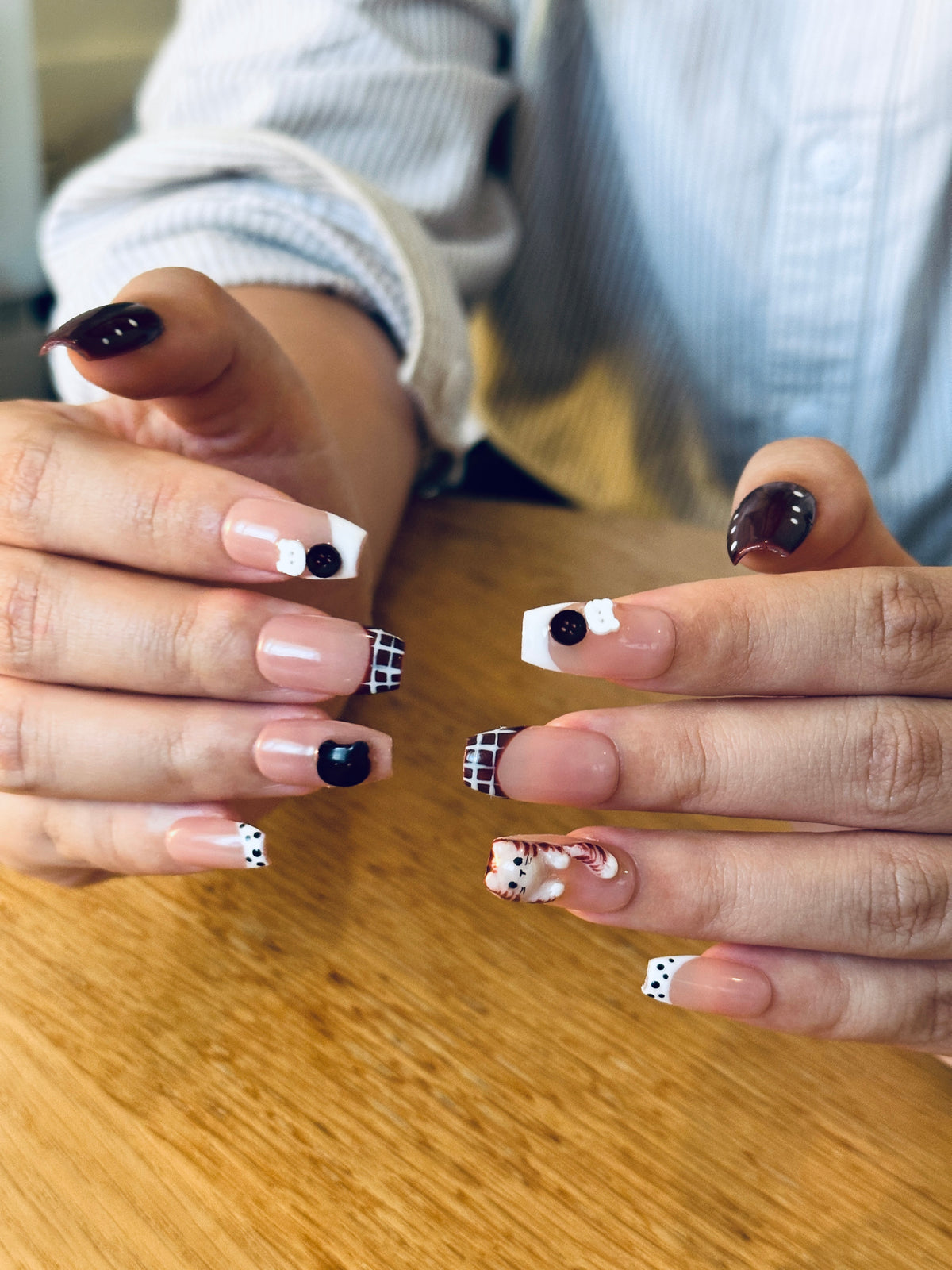 Hands posed on a wooden table displaying coffin press-on nails with dark brown French tips, classic plaid, vintage buttons, and a 3D cat charm
