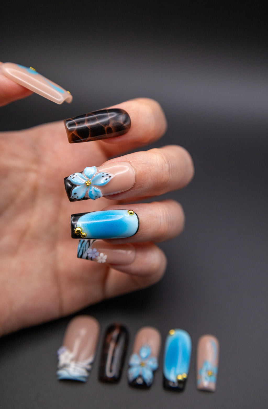 Hand showing glossy blue aura coffin press-on nails adorned with 3D sculpted flowers, tortoise pattern, and metallic accents