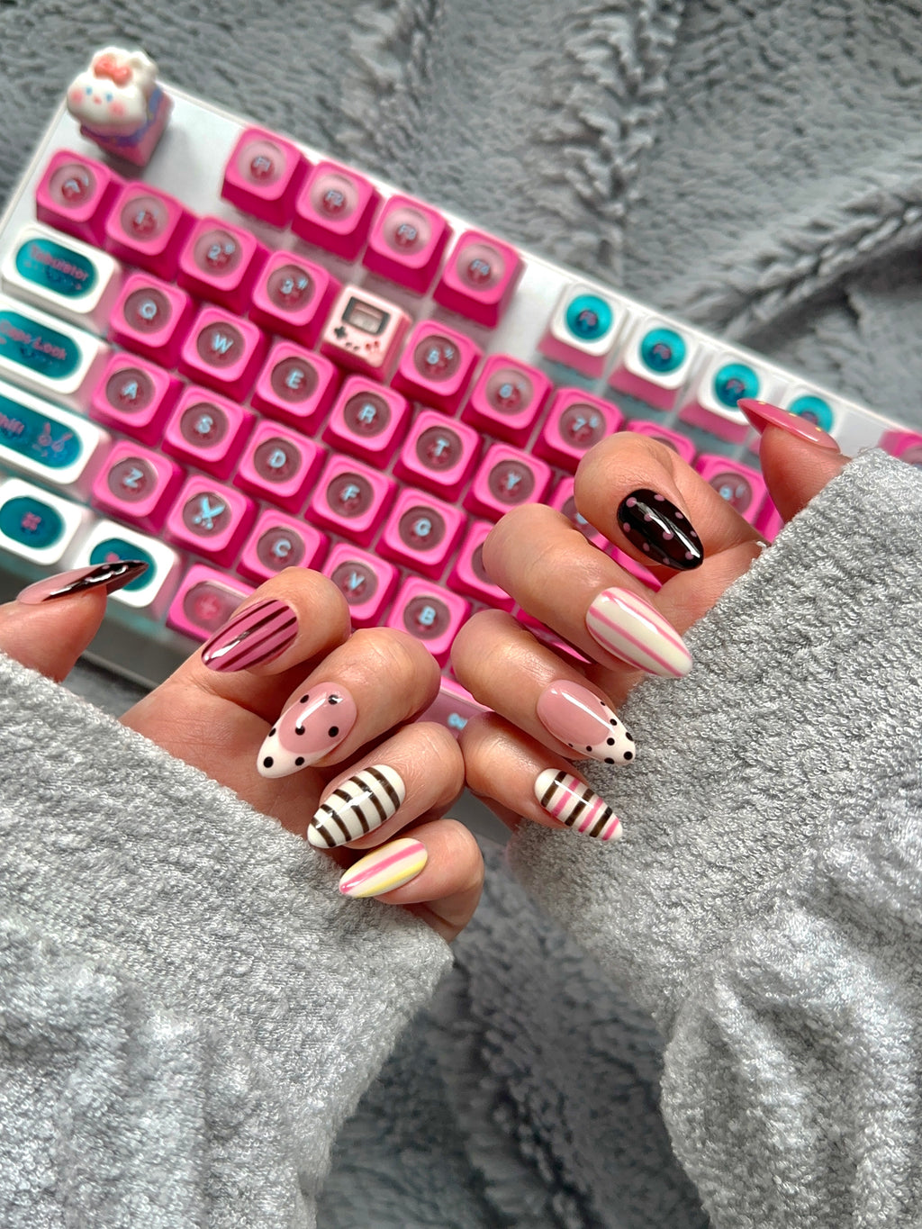 Hands posed in front of a pink mechanical keyboard, displaying oval press-on nails with glossy pink and black polka dots and candy stripe patterns