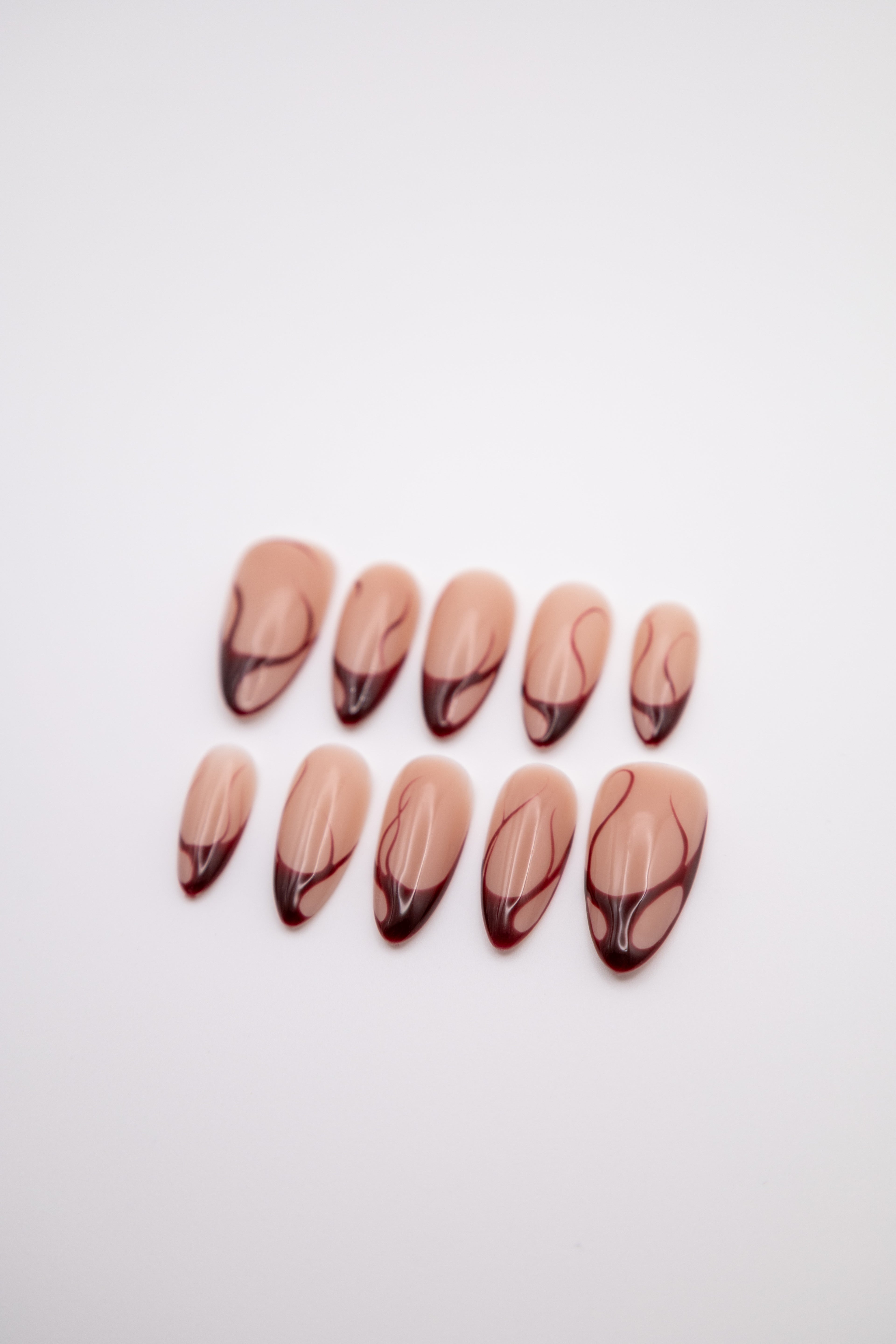 Flatlay of glossy nude almond press-on nails with deep red swirl french tips