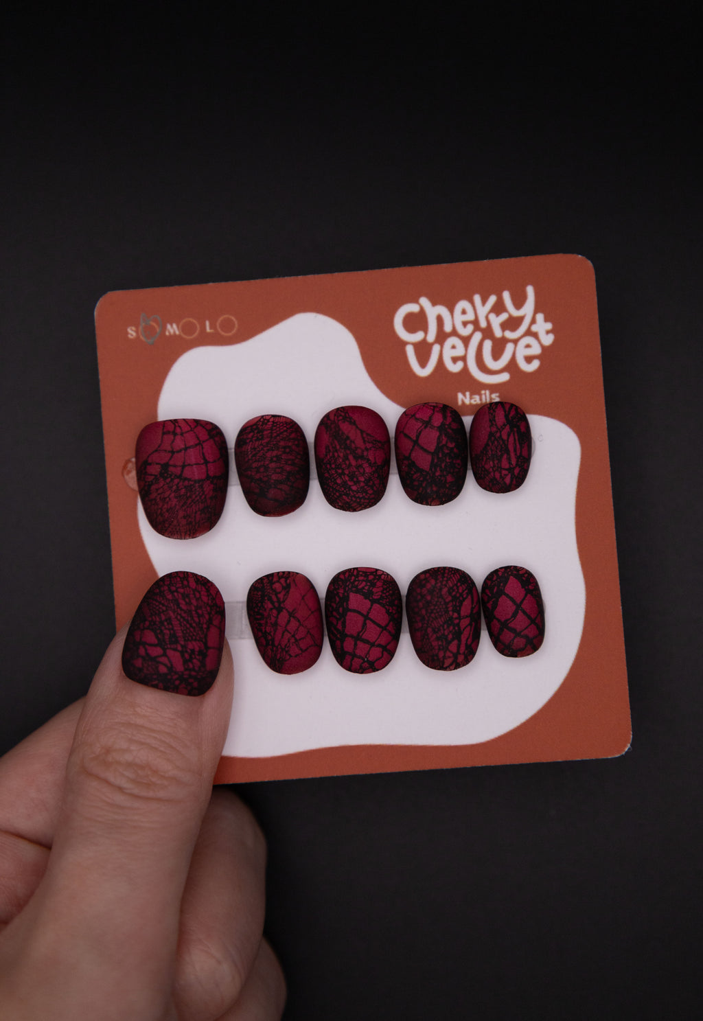 Hand holding CherryVelvet display card showing matte burgundy red press-on nails with intricate black lace patterns