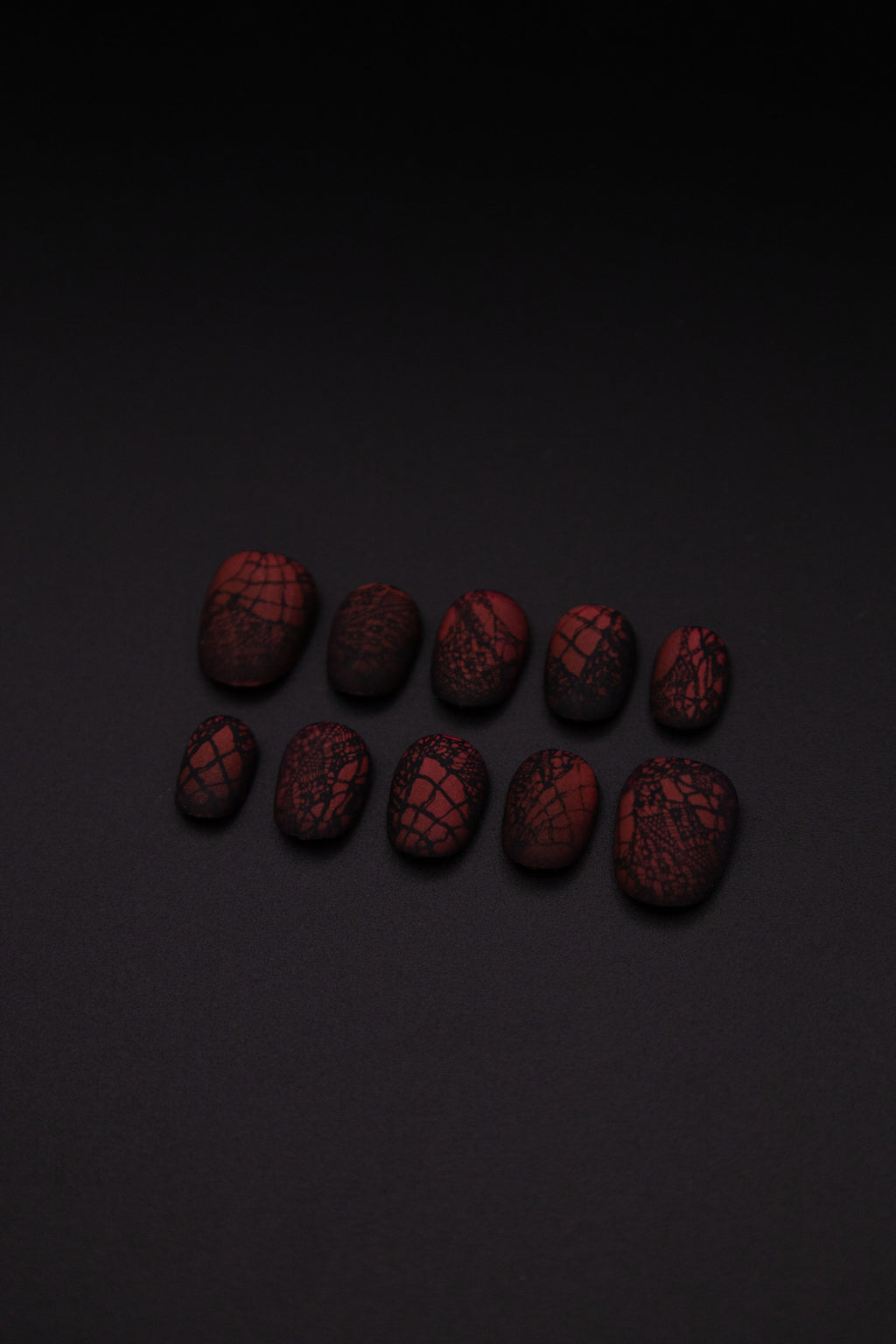 Flatlay of 10 matte burgundy press-on nails with delicate black lace design against a sleek black background