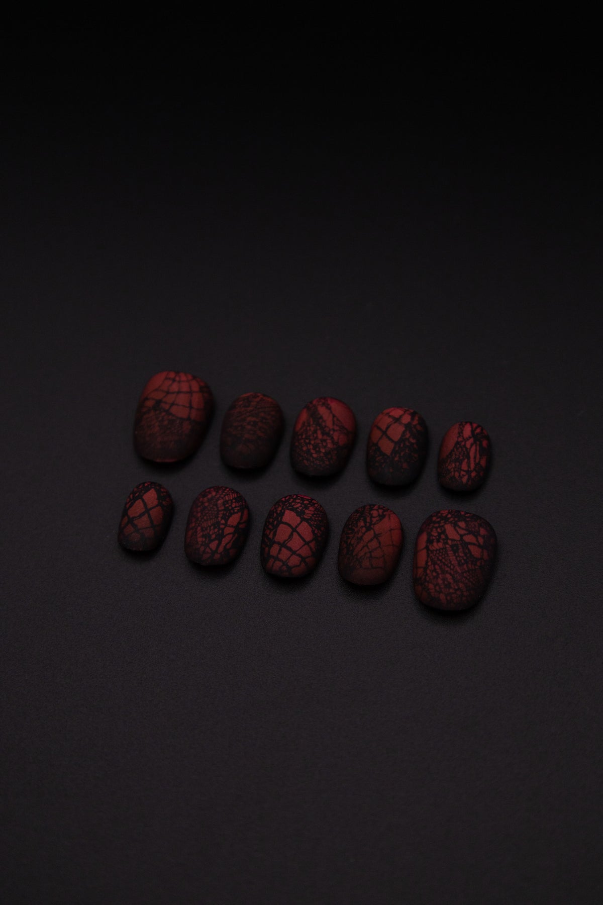 Flatlay of 10 matte burgundy press-on nails with delicate black lace design against a sleek black background