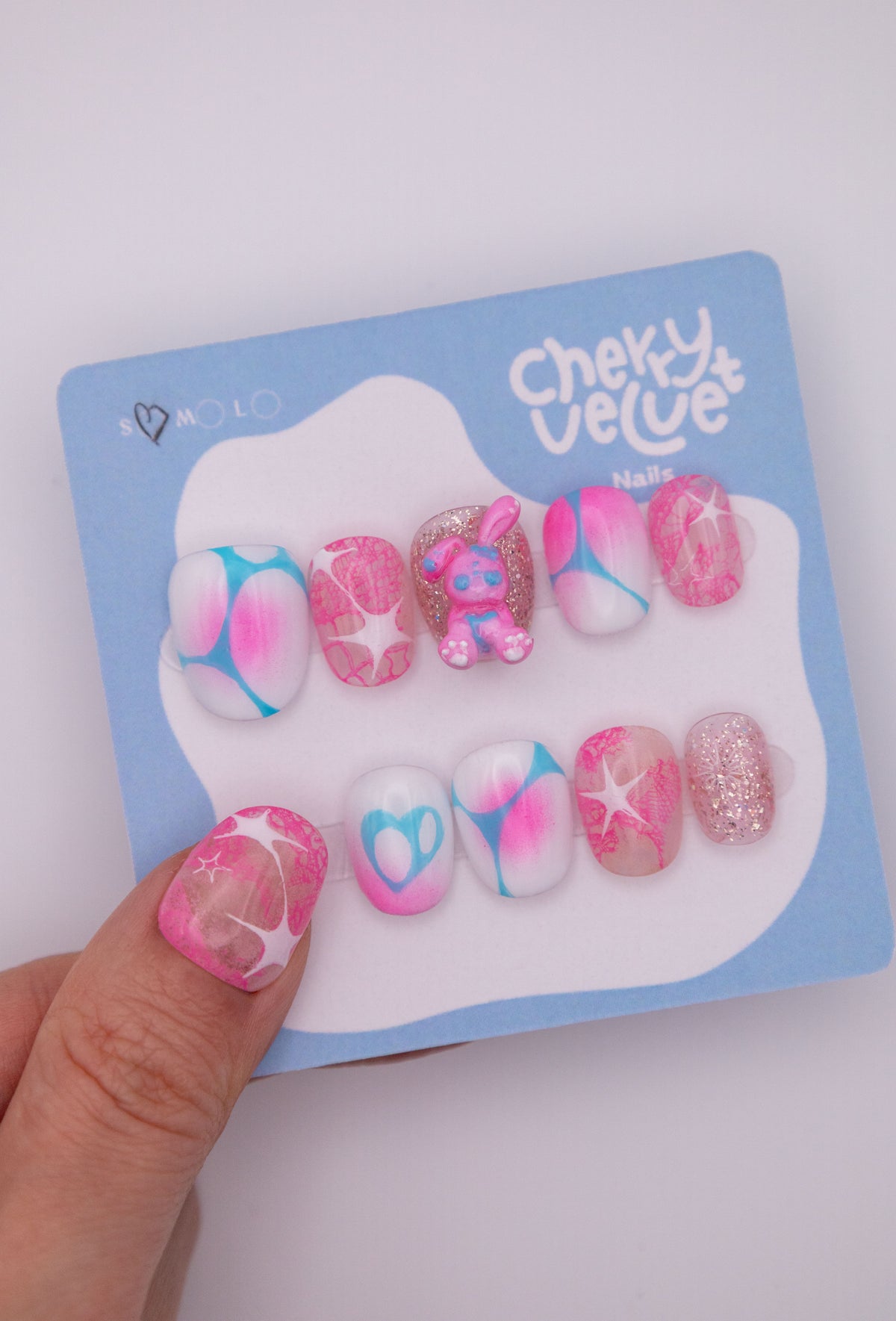 Hand holding blue CherryVelvet display card showing short oval press-on nails in pink and blue Y2K star patterns with a 3D bunny charm