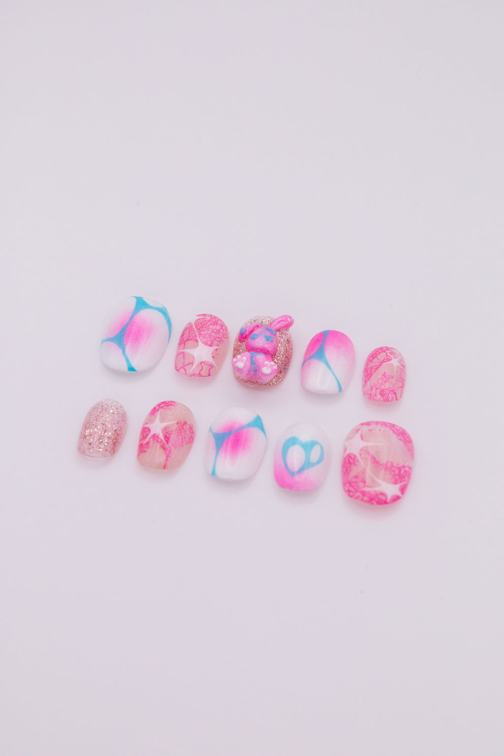 Flatlay of 10 short oval press-on nails featuring vibrant pink and blue aura gradients, Y2K starbursts, glitter, and a cute 3D bunny charm