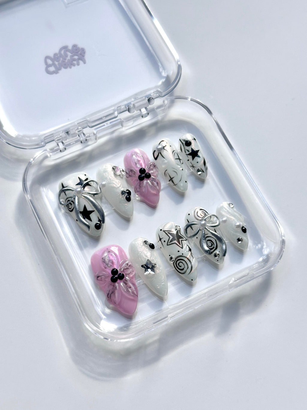 Clear acrylic display box filled with almond press-on nails featuring 3D pink flowers, silver metallic ribbons, Y2K stars, and pearl accents