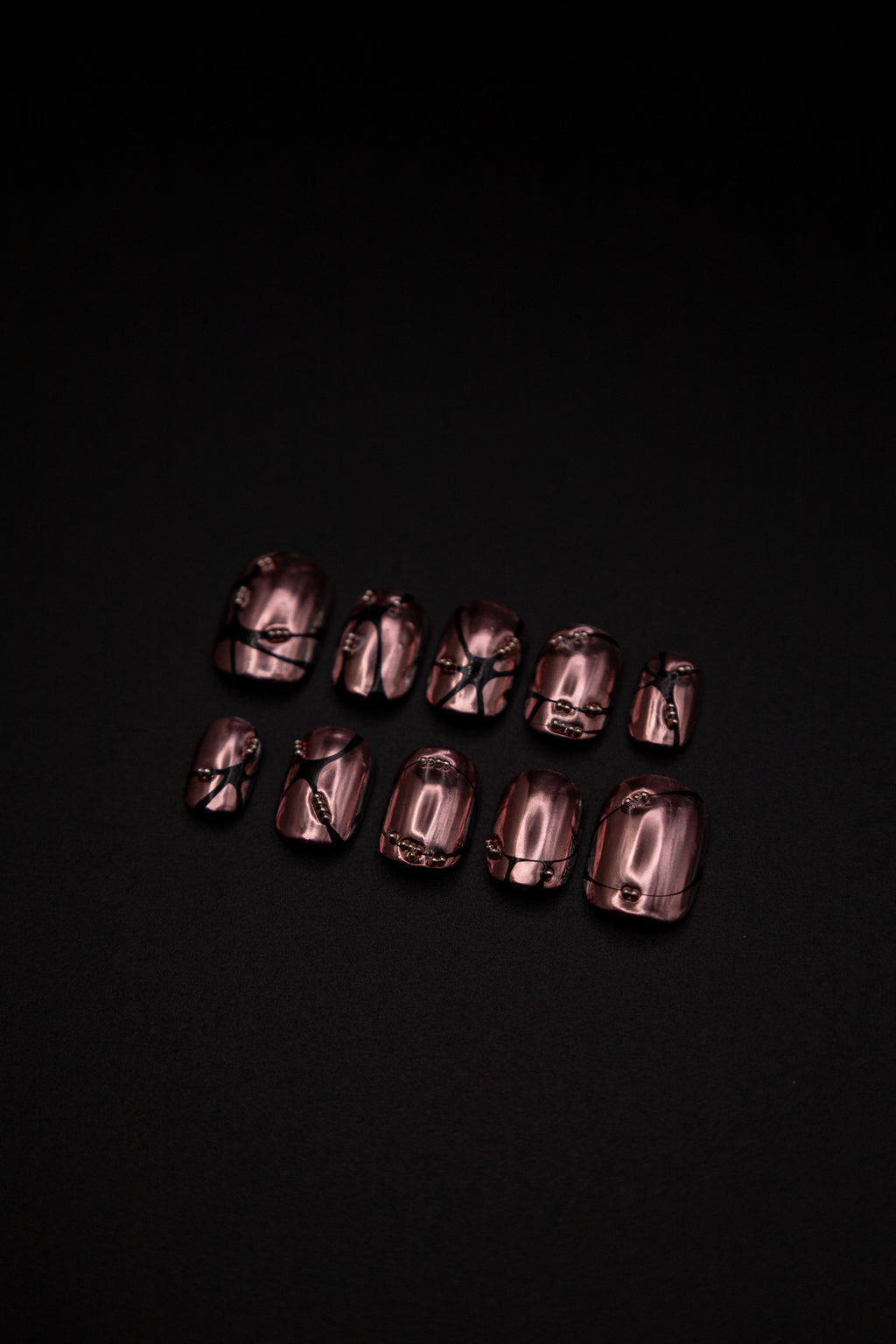 Flatlay of short rose bronze chrome press-on nails featuring 3D black web lines and metallic studs