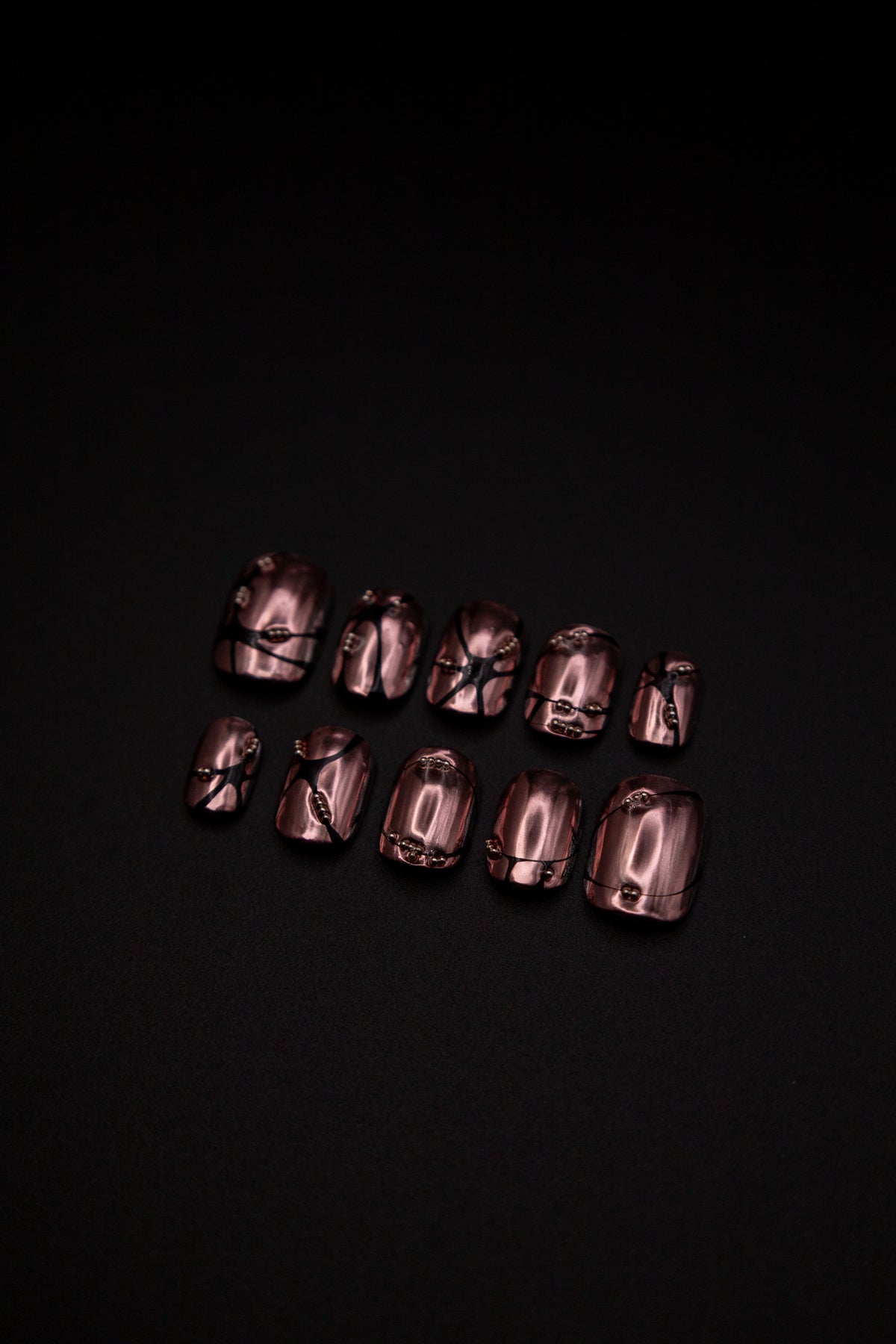 Flatlay of short rose bronze chrome press-on nails featuring 3D black web lines and metallic studs