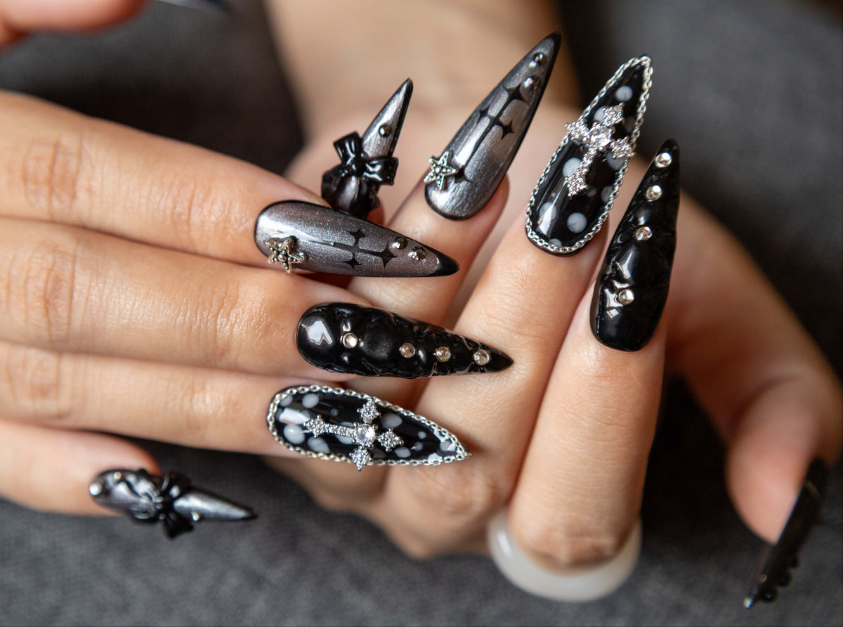 Close-up of hands wearing dark gothic stiletto press-on nails with silver metallic crosses, 3D textured bows, and sparkling black glitter accents