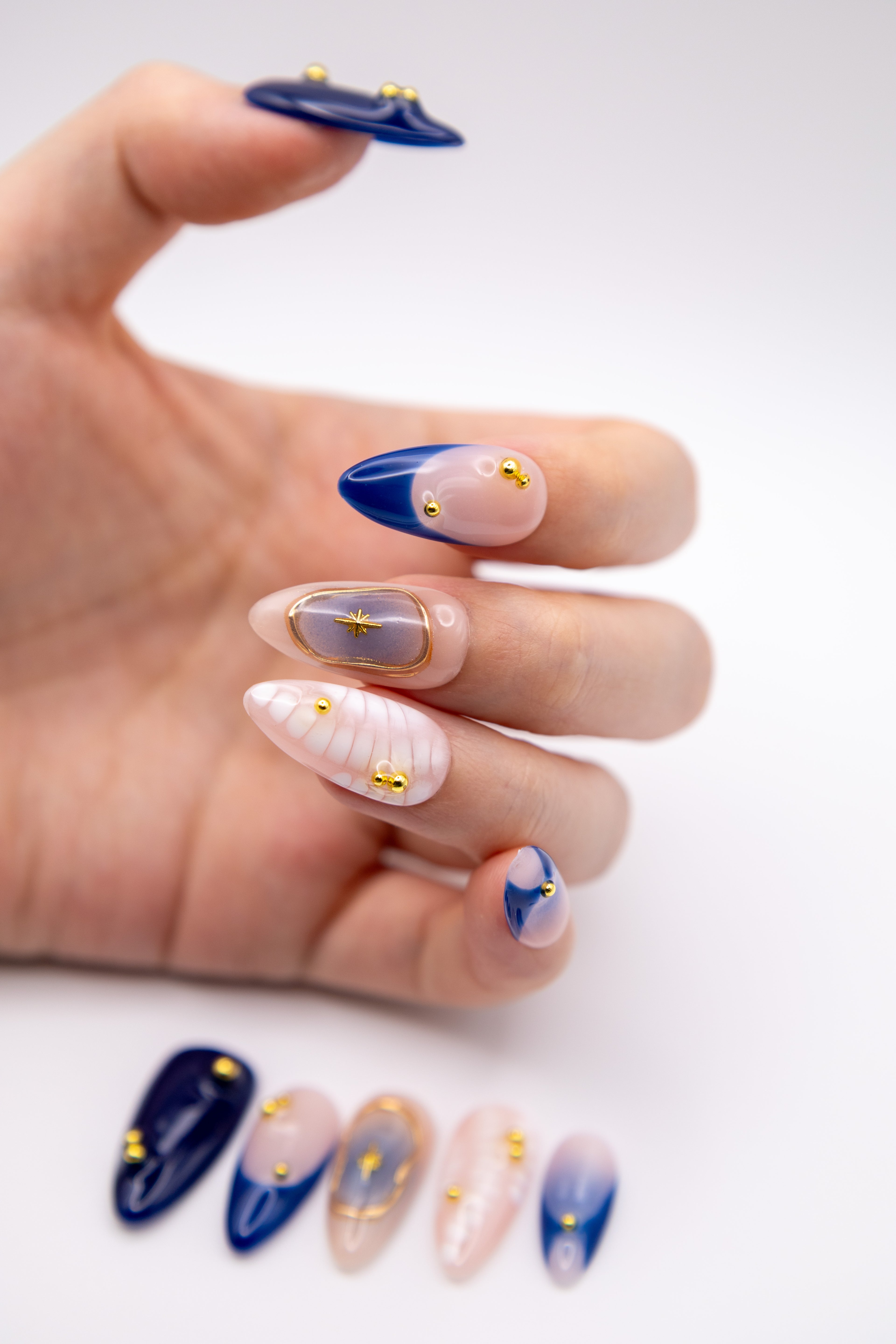 Hand showing glossy deep blue blush press-on nails with 3D relief design, gold starburst, and metal beads