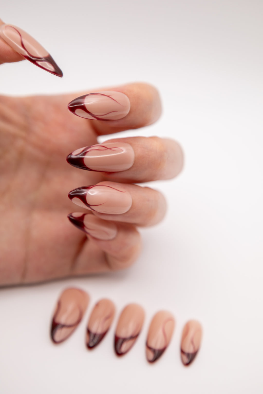 Hand showing glossy nude almond press-on nails featuring elegant red swirling french lines