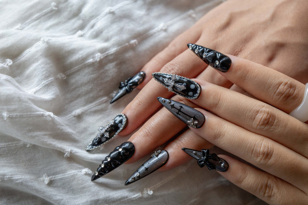 Hand displaying dark gothic press-on nails with silver chain rhinestone cross charms and sparkling black shimmer on a white textured fabric background