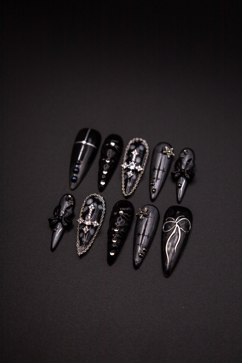 Flatlay of long stiletto press-on nails featuring sparkling black shimmer, silver rhinestone crosses with chain borders, and 3D bow charms