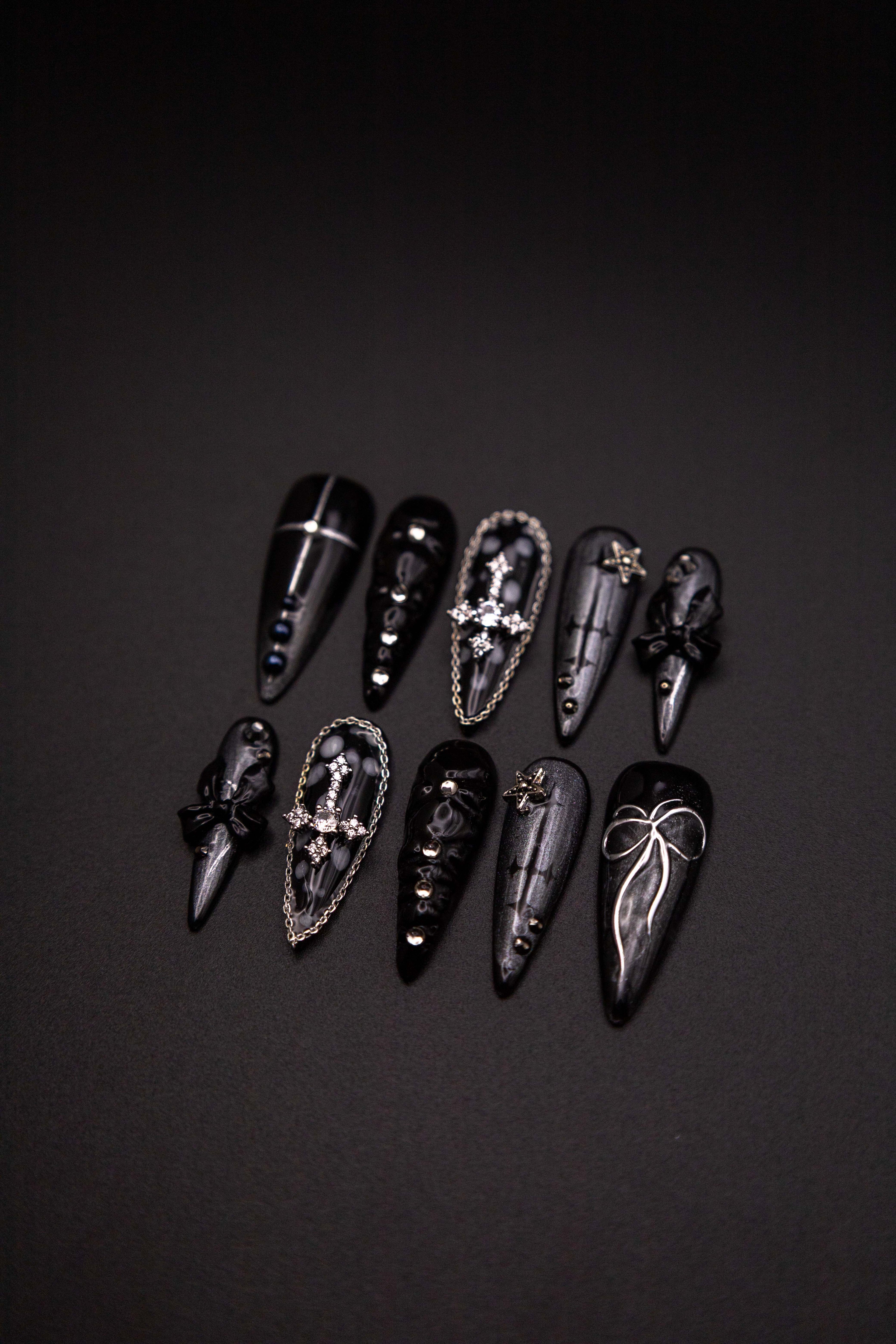 Flatlay of long stiletto press-on nails featuring sparkling black shimmer, silver rhinestone crosses with chain borders, and 3D bow charms