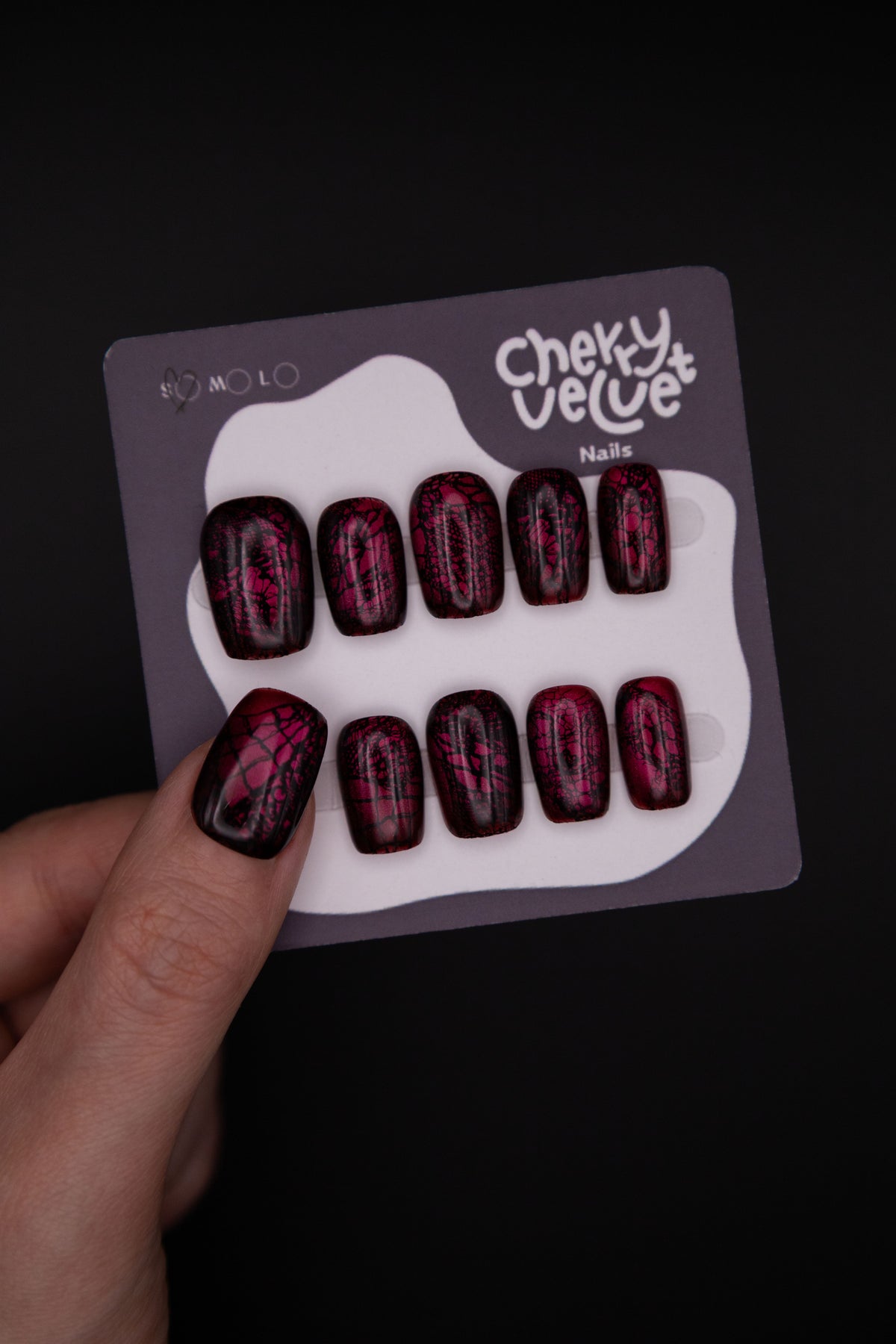 Hand holding CherryVelvet display card showing glossy burgundy press-on nails with shiny black lace overlay