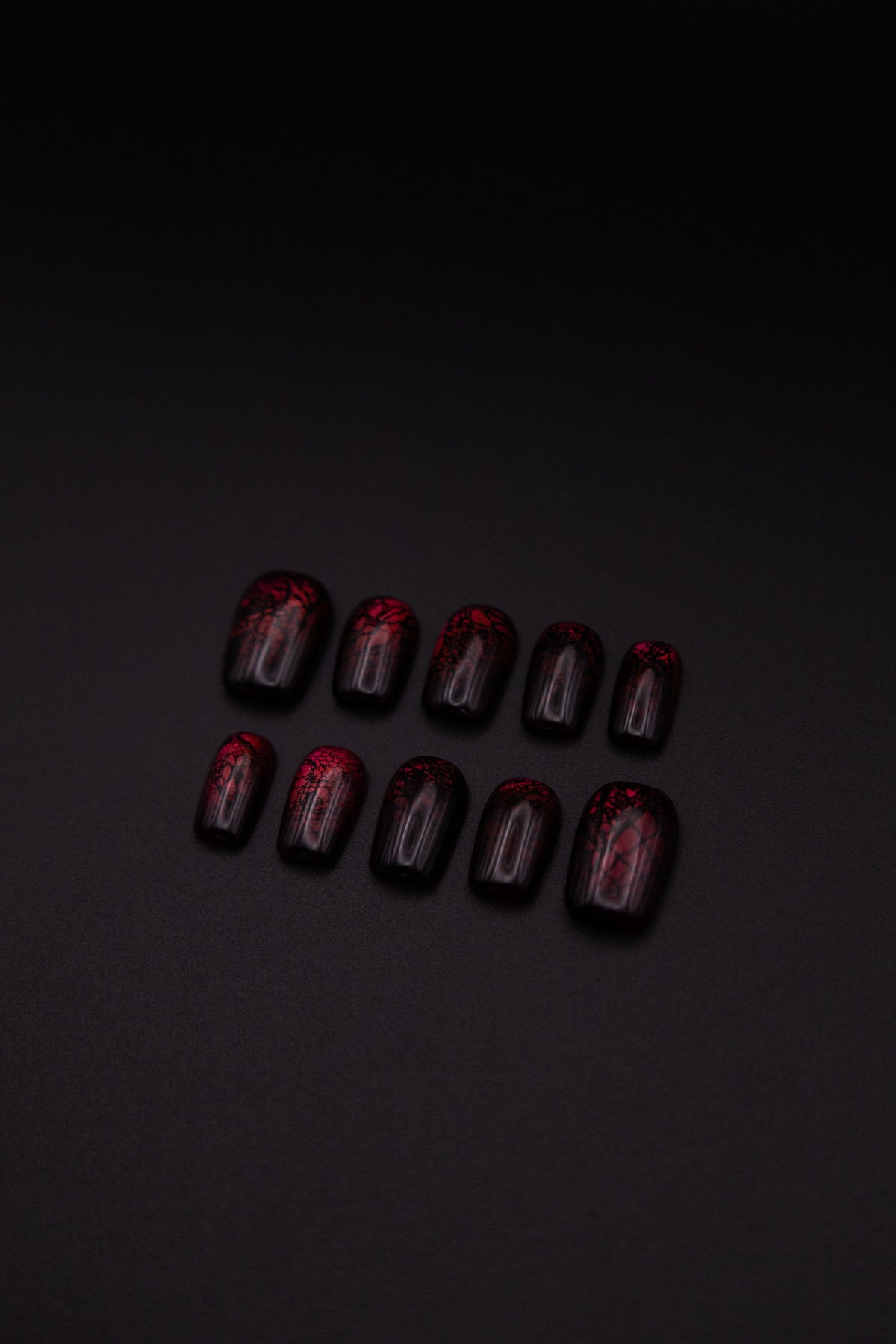 Flatlay of 10 medium squoval glossy burgundy press-on nails with black lace patterns on a black background