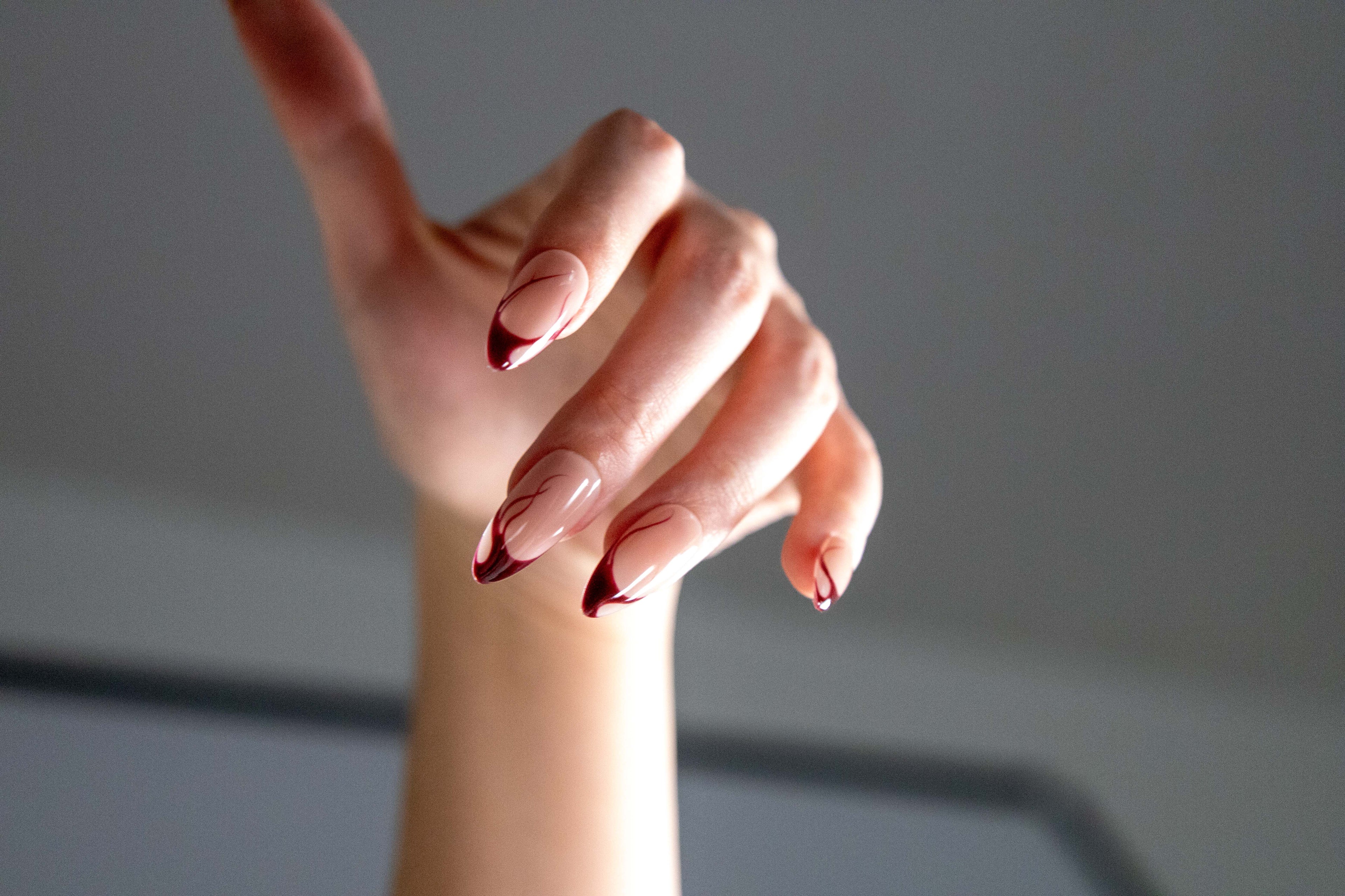 Atmospheric photo of hand wearing burgundy red swirl french almond press-on nails in dramatic lighting