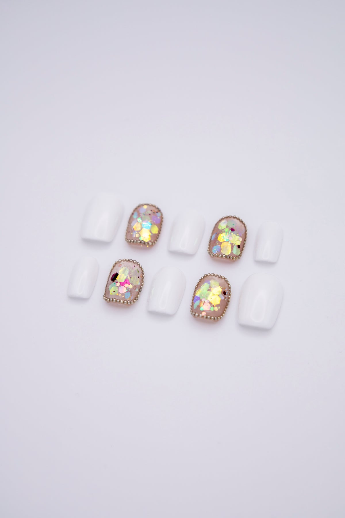 Flatlay of 10 short squoval press-on nails featuring solid milky white nails and iridescent chunky sequins with tiny bead borders