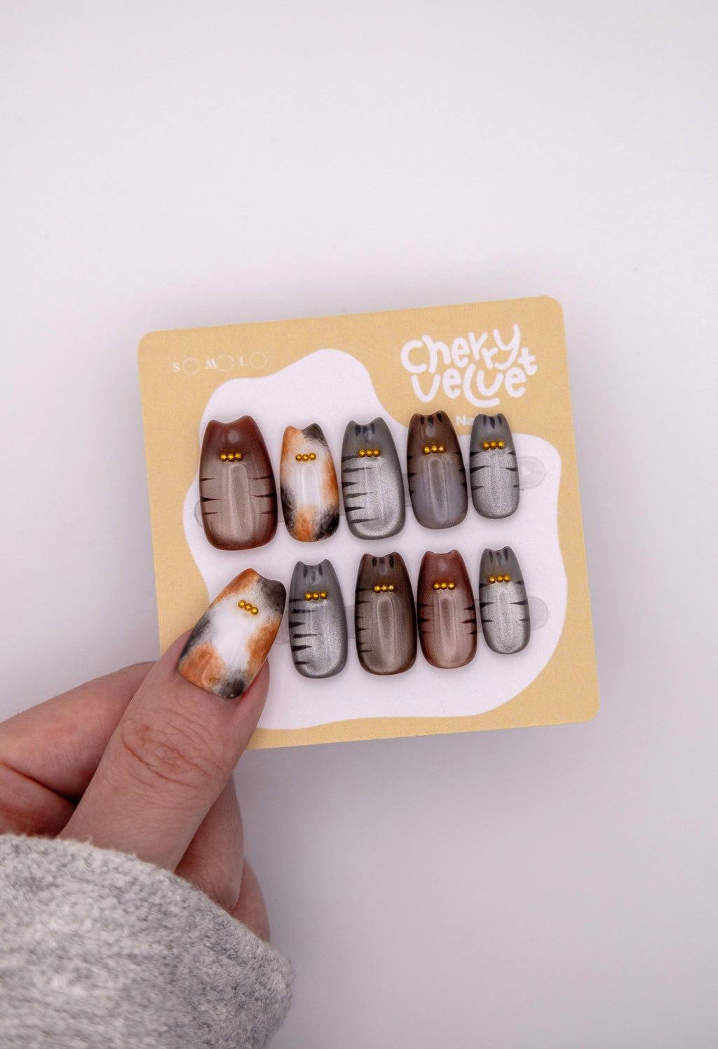 Hand holding CherryVelvet display card showing cat ear shaped press-on nails in calico and tabby patterns with tiny gold beads