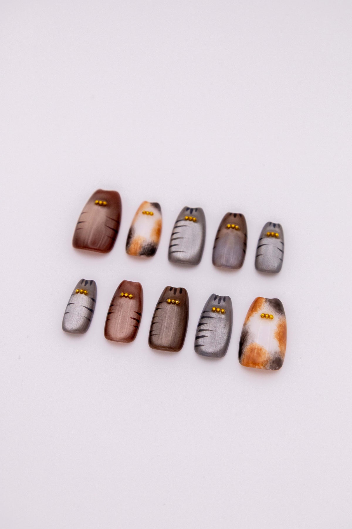 Flatlay of 10 cat ear shaped press-on nails with hand-painted calico patches, tabby stripes, and gold bead accents on a clean background