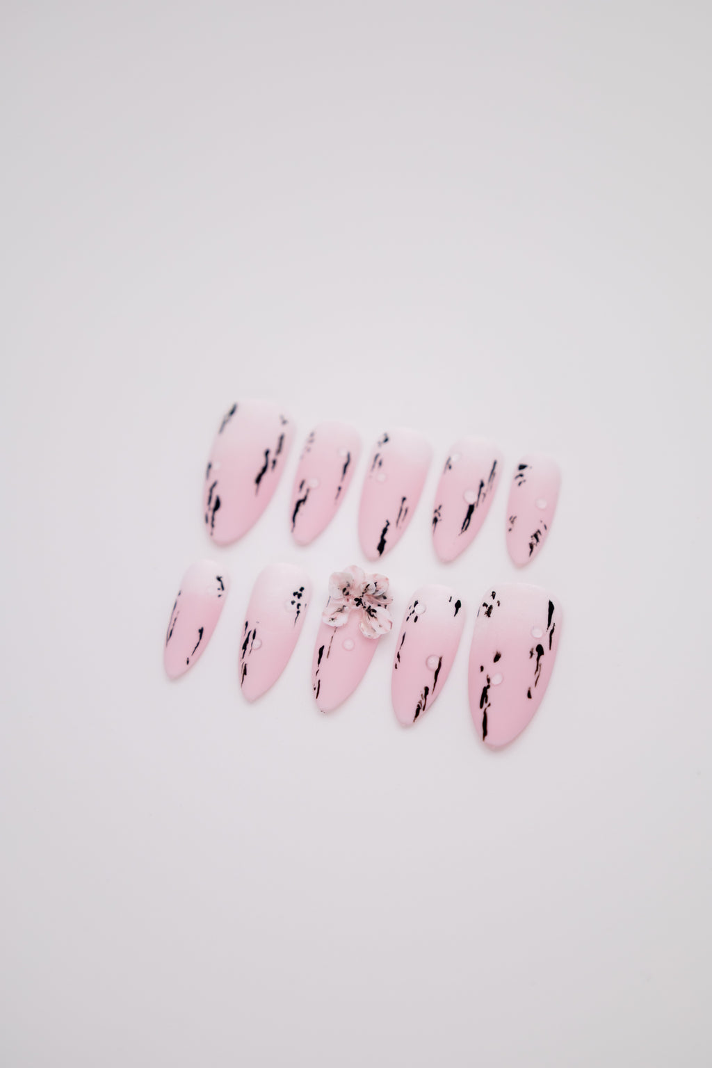 Flatlay of matte pink white gradient almond press-on nails featuring black ink strokes and a 3D floral charm