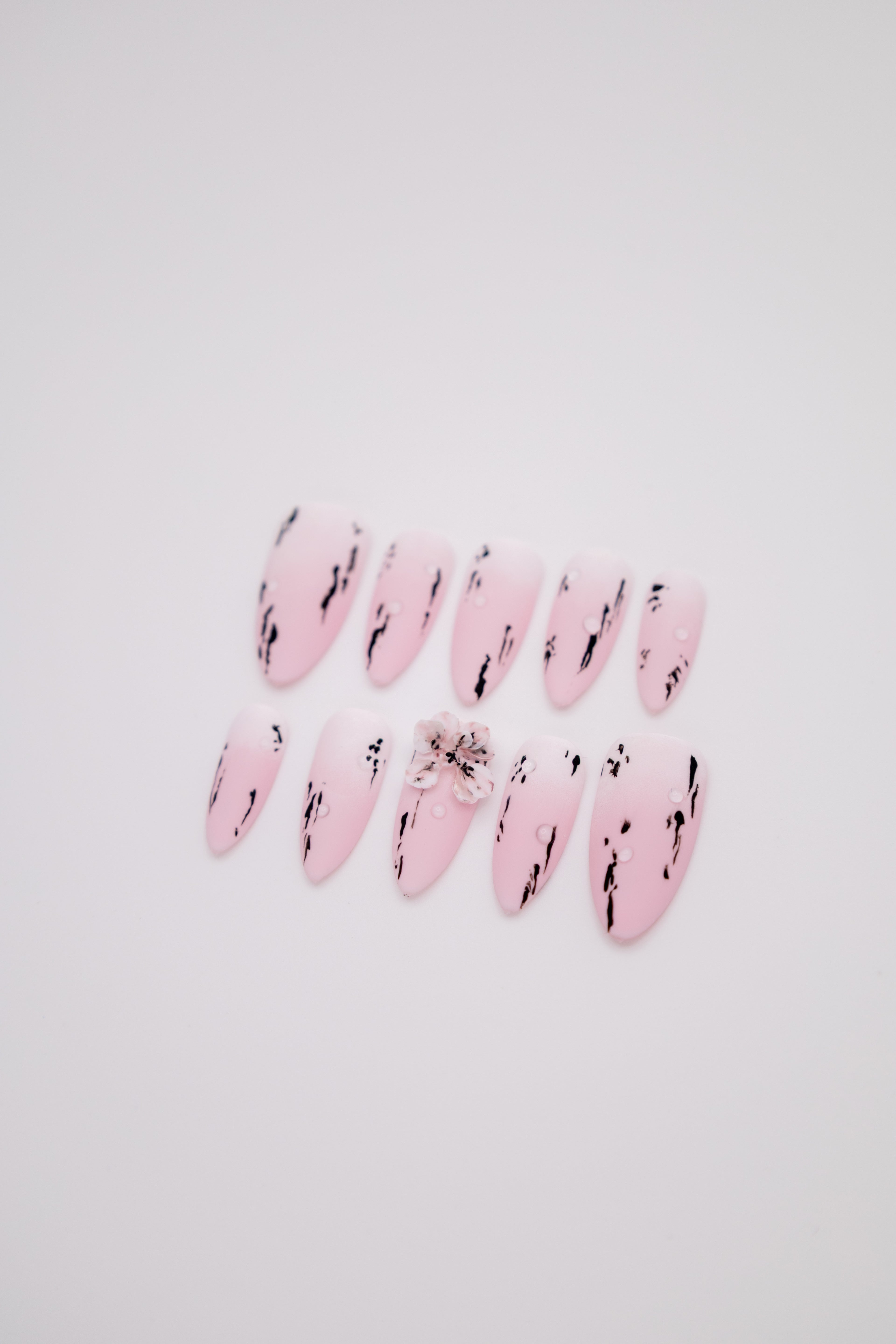 Flatlay of matte pink white gradient almond press-on nails featuring black ink strokes and a 3D floral charm