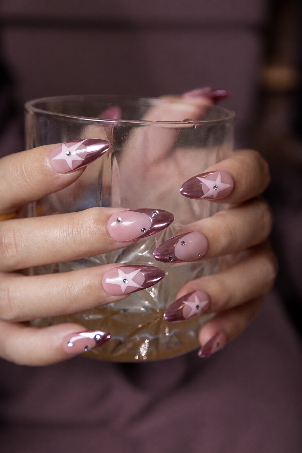 Hands holding a glass, displaying almond press-on nails with metallic pink chrome French tips, hollow star designs, and sparkling rhinestones