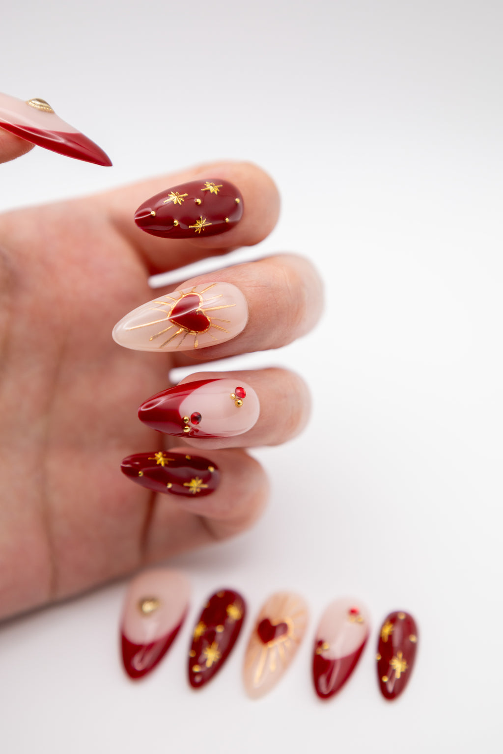 Hand showing glossy red almond press-on nails with radiant gold heart art, starbursts, and red gem accents