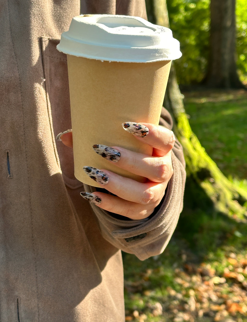Hand wearing Sepia Dapple almond press-on nails holding a paper coffee cup outdoors, showing tortoise pattern and artistic brushstroke designs in natural sunlight