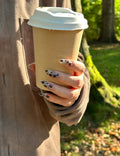 Hand wearing Sepia Dapple almond press-on nails holding a paper coffee cup outdoors, showing tortoise pattern and artistic brushstroke designs in natural sunlight