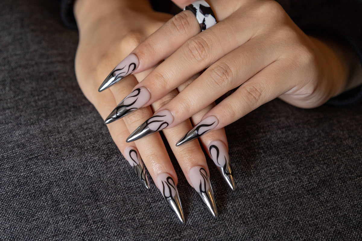 Both hands crossed displaying glossy nude press-on nails featuring silver chrome metallic French tips and black cyber flame designs