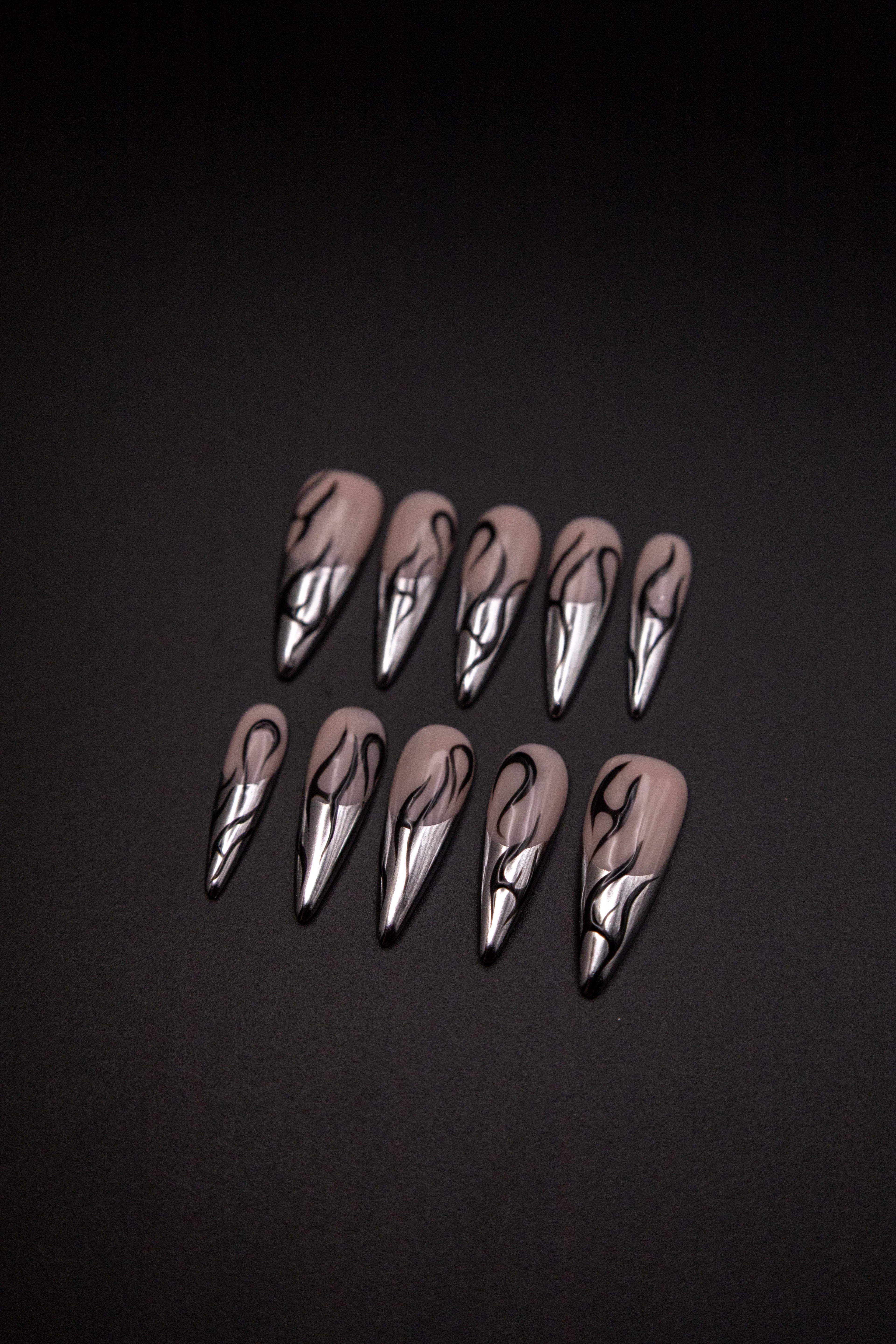 Flatlay of long almond press-on nails featuring liquid silver chrome French tips and black tribal flame line art