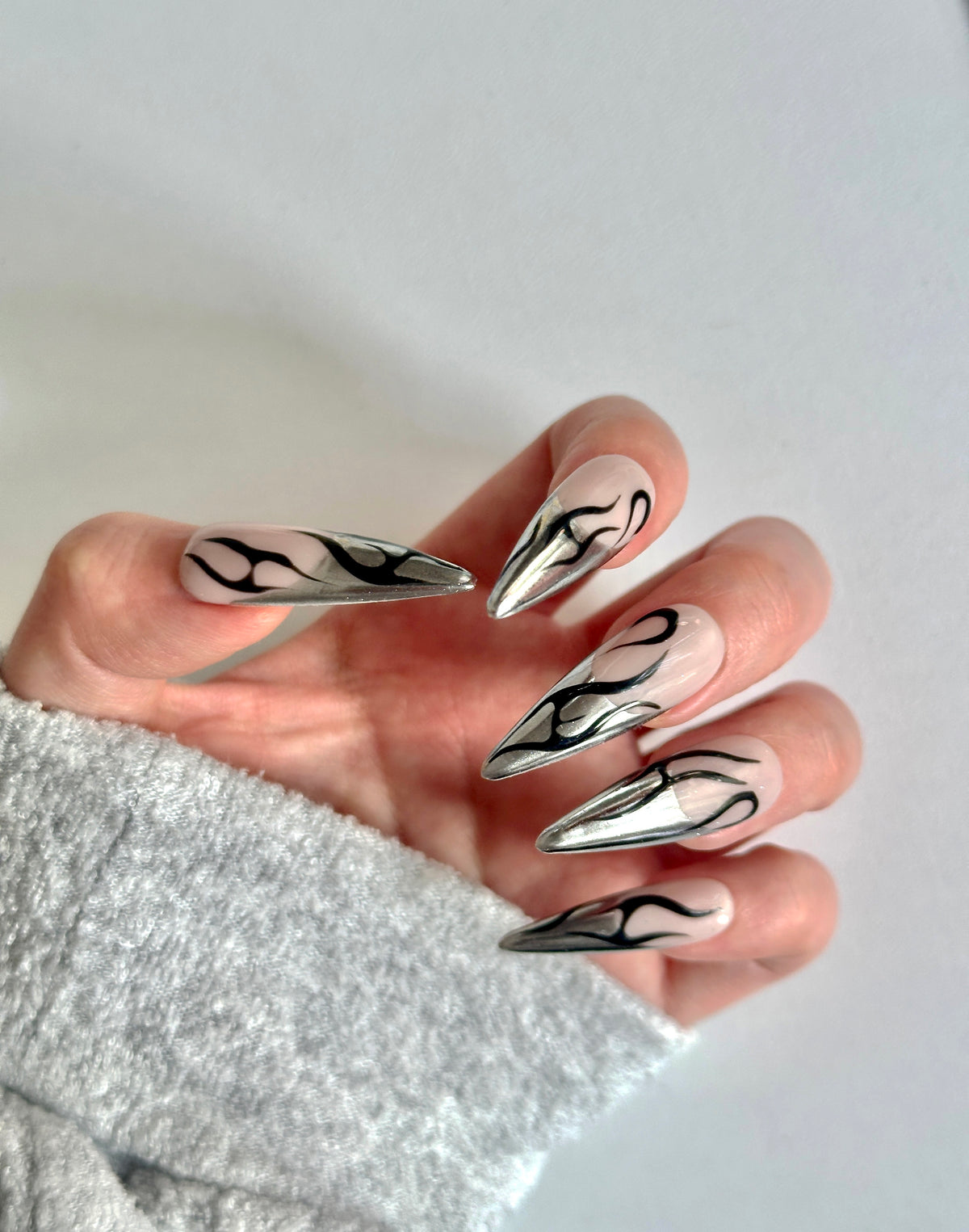 Close-up of a hand with a grey sweater sleeve, displaying long almond press-on nails with metallic silver chrome French tips and black tribal flame line art