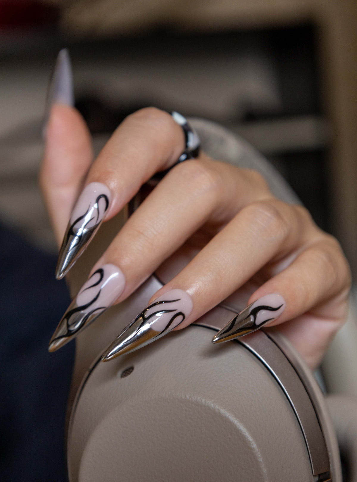 Close-up of a hand holding headphones, showcasing long stiletto press-on nails with metallic silver chrome French tips and black flame art