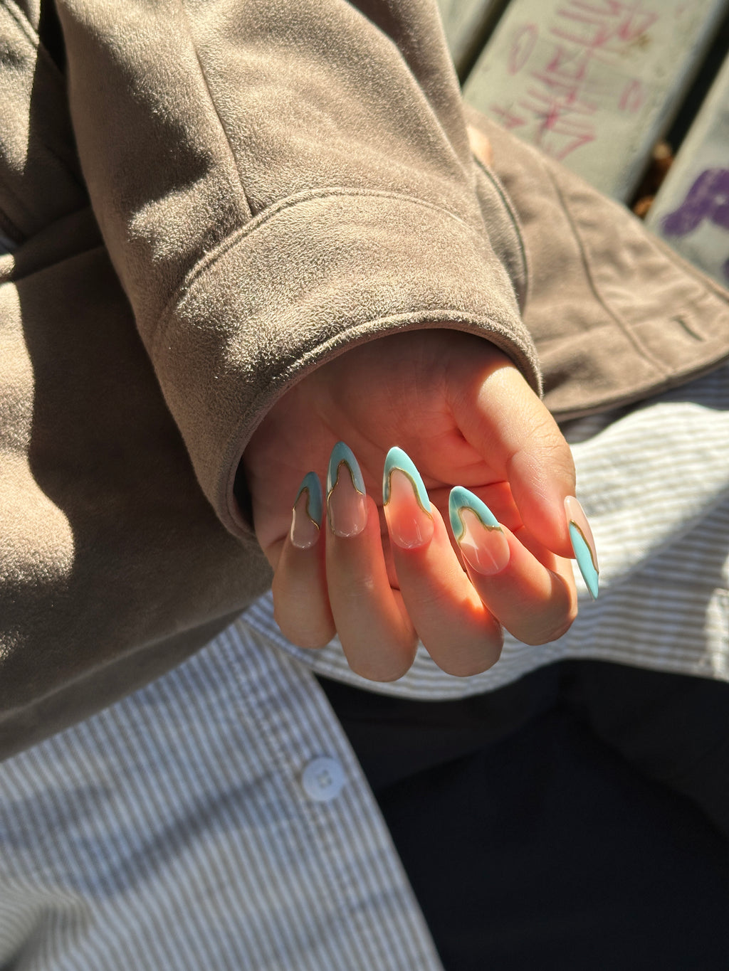 Close-up of a hand in natural sunlight, displaying oval press-on nails featuring sky blue wavy French tips outlined in metallic gold