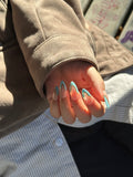 Close-up of a hand in natural sunlight, displaying oval press-on nails featuring sky blue wavy French tips outlined in metallic gold